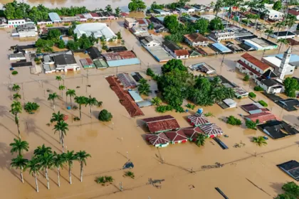 Rua de cidade alagada após fortes chuvas, com casas e veículos parcialmente submersos pela enchente.