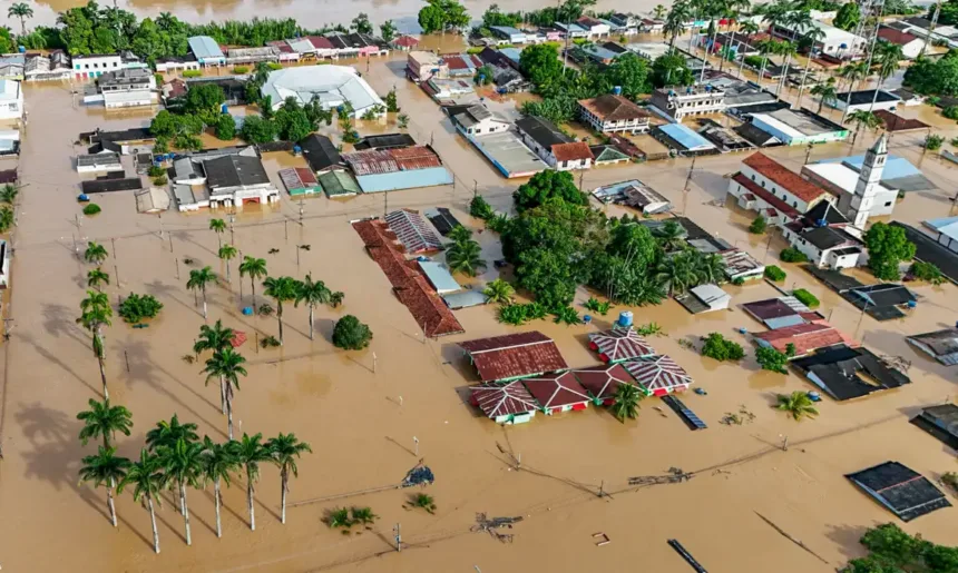 Rua de cidade alagada após fortes chuvas, com casas e veículos parcialmente submersos pela enchente.